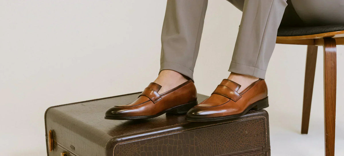 Person wearing polished brown penny loafers while standing on a vintage suitcase indoors