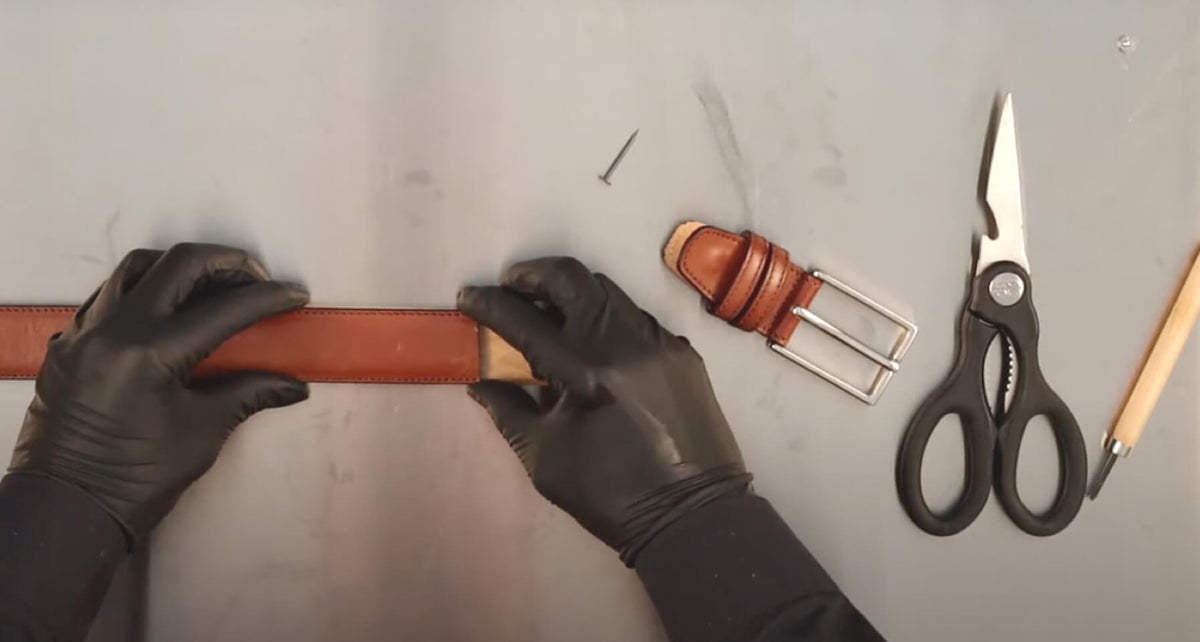 Gloved hands position a brown leather belt beside its buckle, scissors, and pencil, showing how to cut the belt for a custom fit