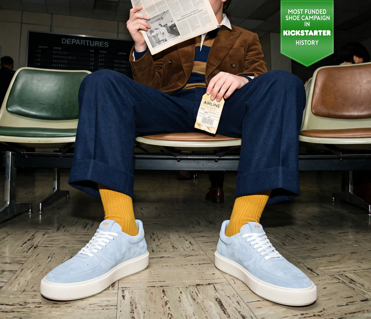 Man sitting in waiting room wearing men's light blue Italian suede leather lace-up sneakers with white rubber soles and mustard color socks with navy trousers and a brown jacket.
