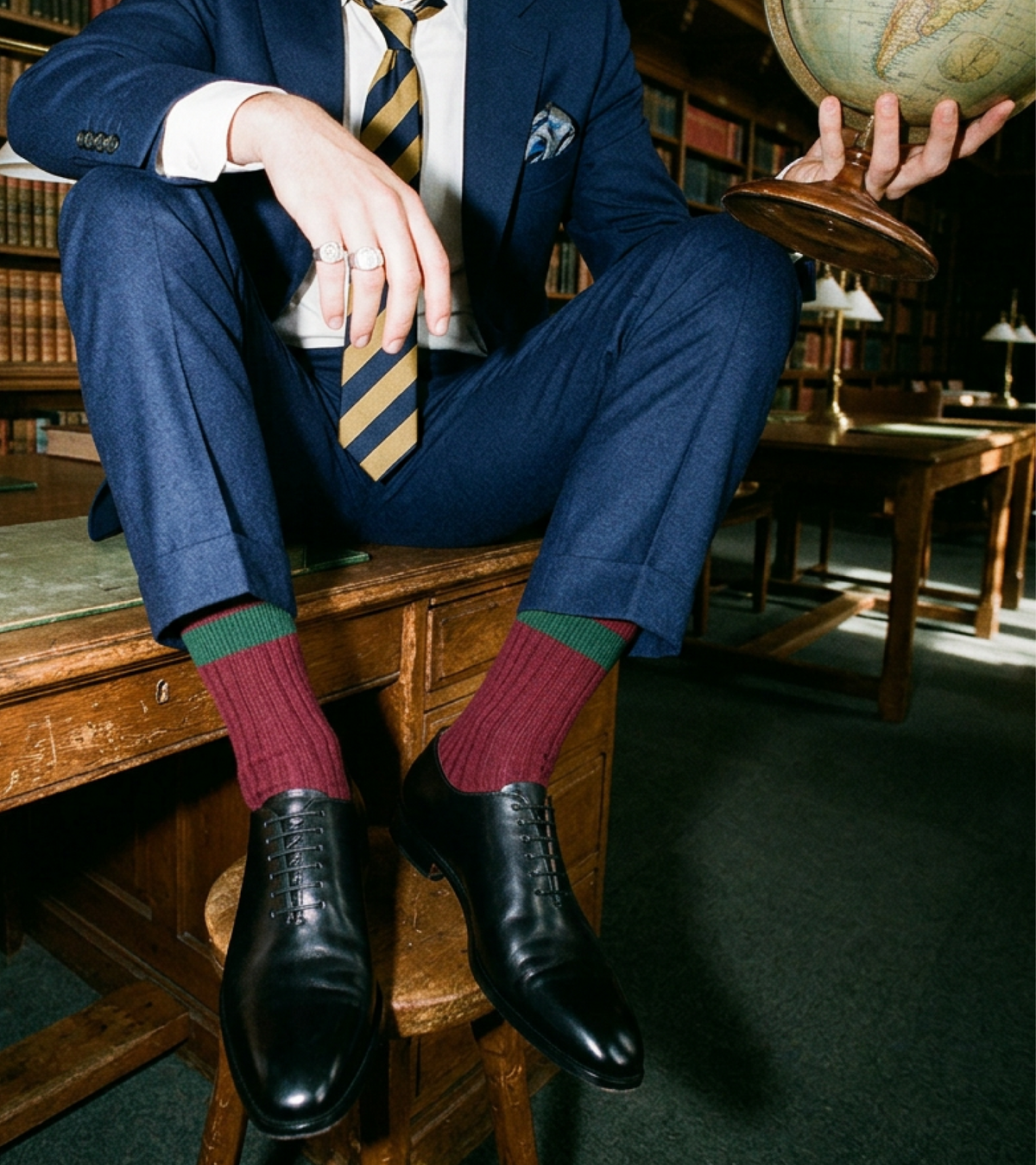 A man in a navy blue suit sits on a wooden desk in a library, wearing burgundy and green striped socks with men's black Italian leather dress shoes. He holds a glass and a globe, with bookshelves and study tables in the background.