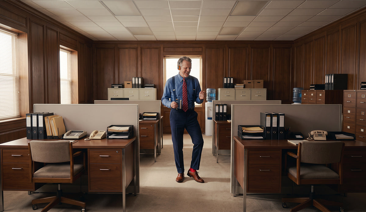 A man in a blue dress shirt, red tie, and navy trousers stands in the middle of a vintage-style wooden office cubicle room, holding a glass of water. Wearing men's Italian brown leather lace-up oxford dress shoes.