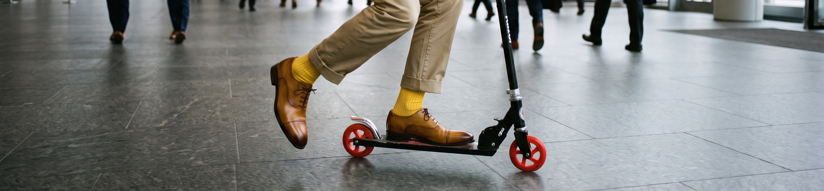 A close-up lifestyle shot of a person's legs wearing tan trousers, men's Italian brown leather lace-up oxford dress shoes, and bright yellow socks while riding a black scooter with red wheels in a modern indoor hallway.