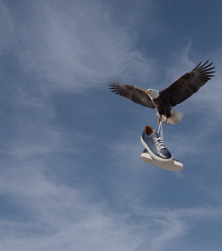 An eagle in flight holds Italian blue leather dress senakers in its talons against a blue sky with wispy clouds.