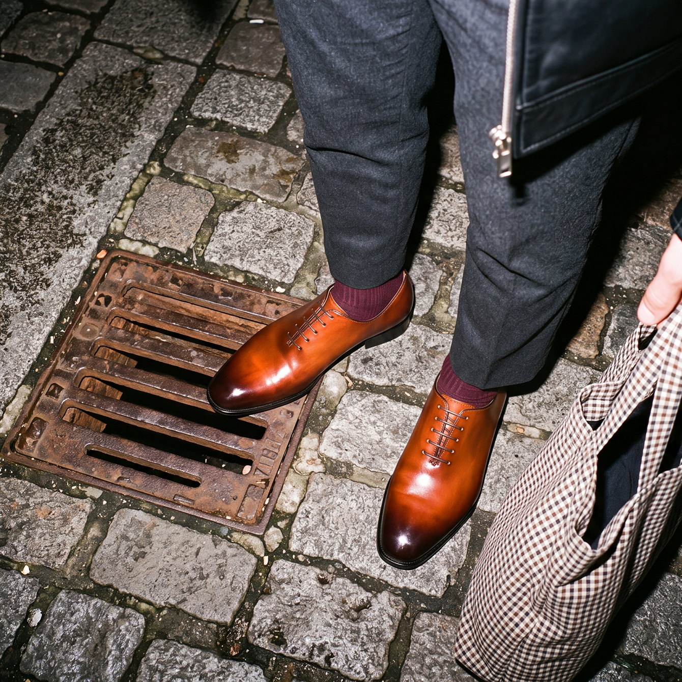 A person wearing dark gray trousers and shiny men's brown Italian leather oxford dress shoes with burgundy socks, standing on a cobblestone street next to a brown and beige houndstooth handbag.