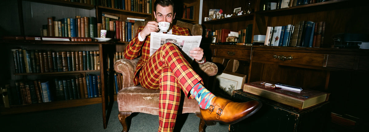 Man sitting in library reading the newspaper wearing a plaid suit and men's caramel brown Italian leather double buckle monk strap dress shoes while sipping coffee.