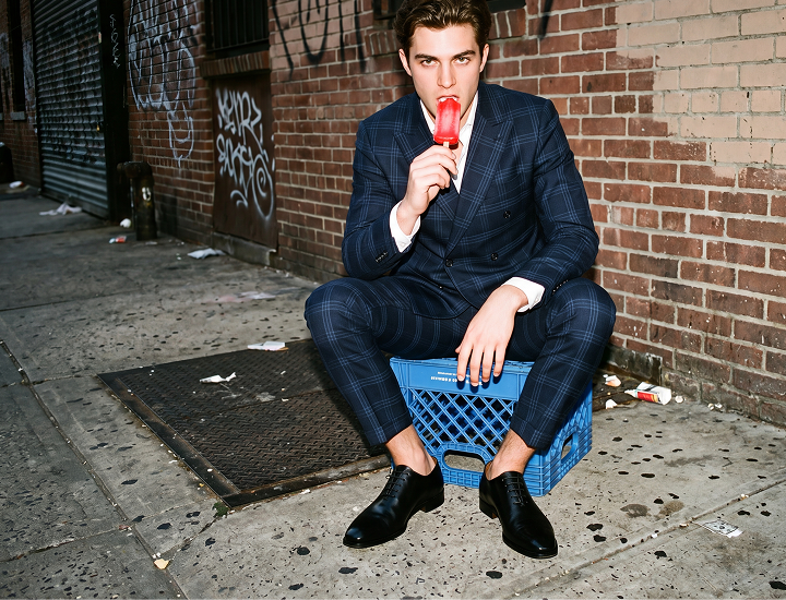 A man wearing men's black Italian leather wholecut lace-up dress shoes in a dark navy plaid suit sits on a blue crate on an urban sidewalk, holding a red popsicle against a brick wall background.