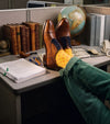 A person's legs and feet resting on a gray office cubicle desk, wearing men's brown Italian leather oxford shoes with yellow patterned socks and green corduroy pants. The desk is cluttered with office items including a globe, binders, books, and papers.