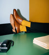 A person's legs crossed with men's brown Italian leather oxford brogue shoes and mustard yellow socks resting on a green desk, with a black telephone and beige folders in the foreground.