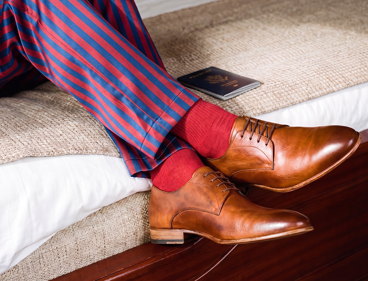 A close-up lifestyle shot of a person's legs crossed on a bed, wearing red and blue striped trousers, red knit socks, and men's Italian brown leather oxford lace-up dress shoes. A passport lies on the bedspread nearby.